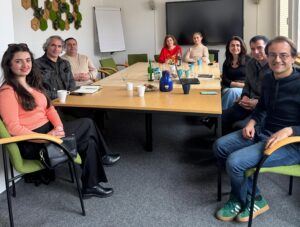 Personen sitzen um einen Besprechungstisch und blicken in die Kamera