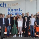 Eine Gruppe von Menschen in die Kamera schauend, darunter Robin Korte, Verena Schäffer, Silke Koch, die eine Urkunde in die Kamera hält. Im Hintergrund der Schriftzug "Es läuft... Kanal-technik"